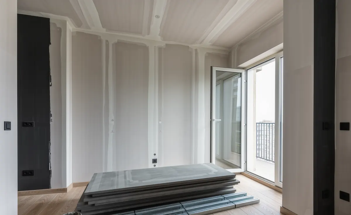 Placas de pladur apiladas junto a perfiles metálicos y un cubo de pasta en el suelo de un piso en obra