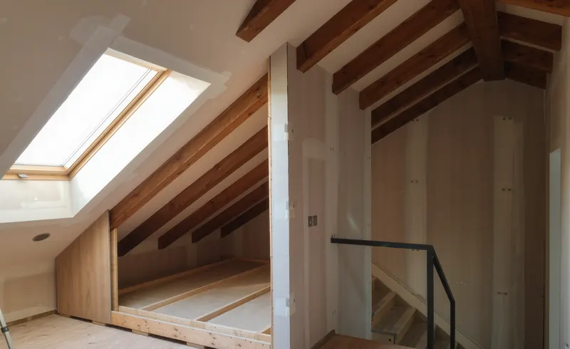 Planta bajo cubierta en obra con vigas de madera vistas, ventana de tejado y un tabique de pladur dividiendo el espacio